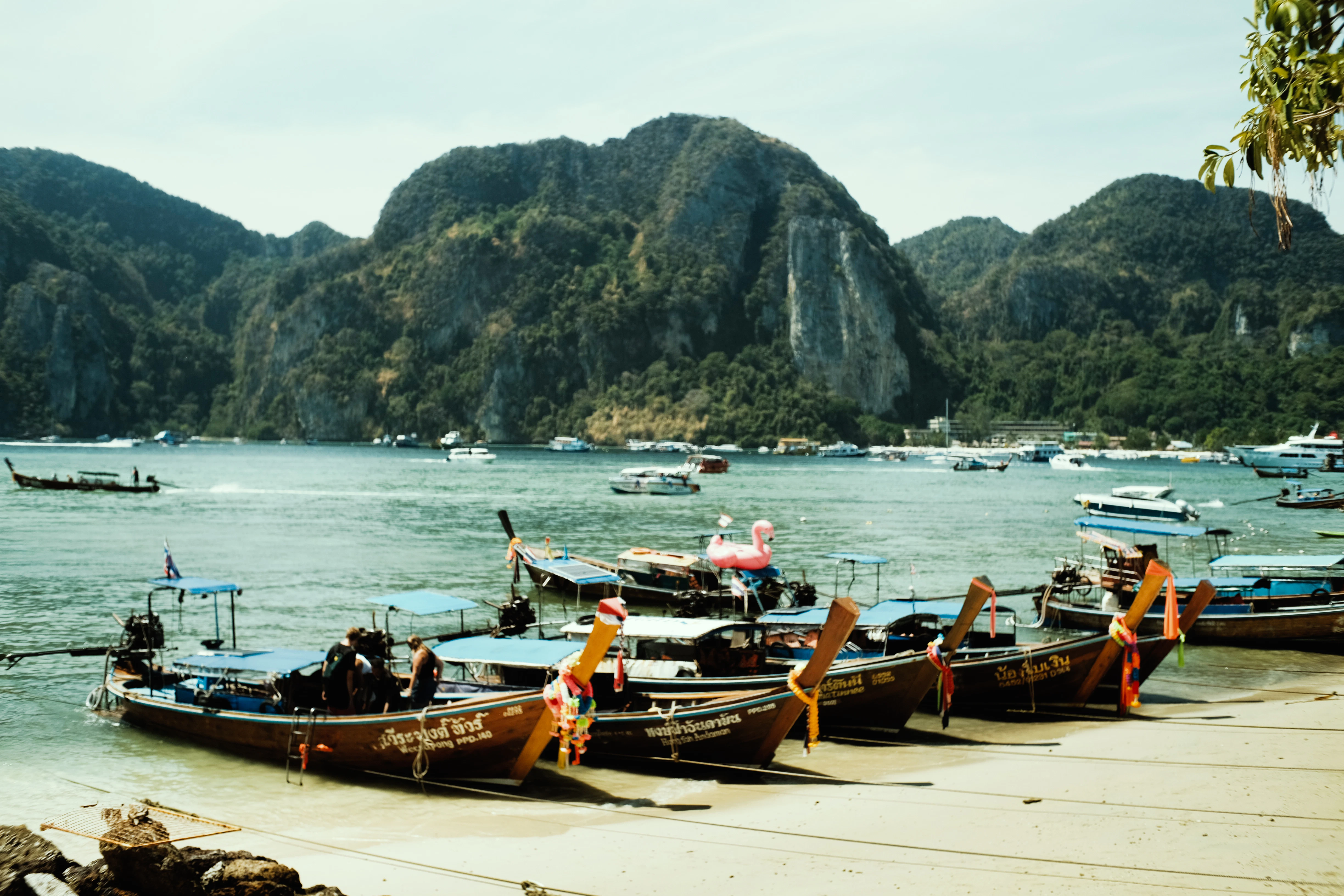 Longtail boats line a sandy beach with mountains behind.