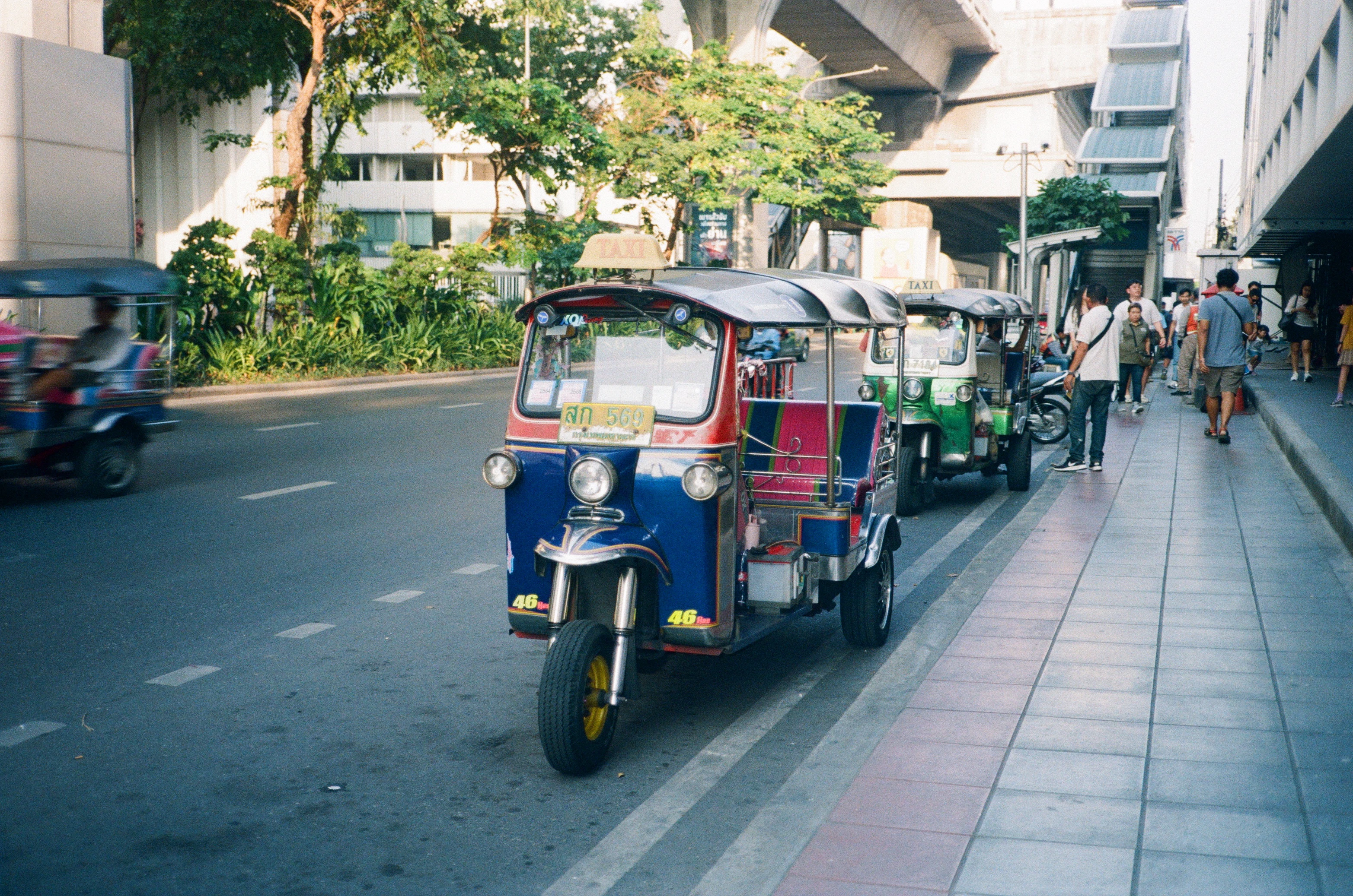 A colorful tuk tuk driving down a street next to tall buildings