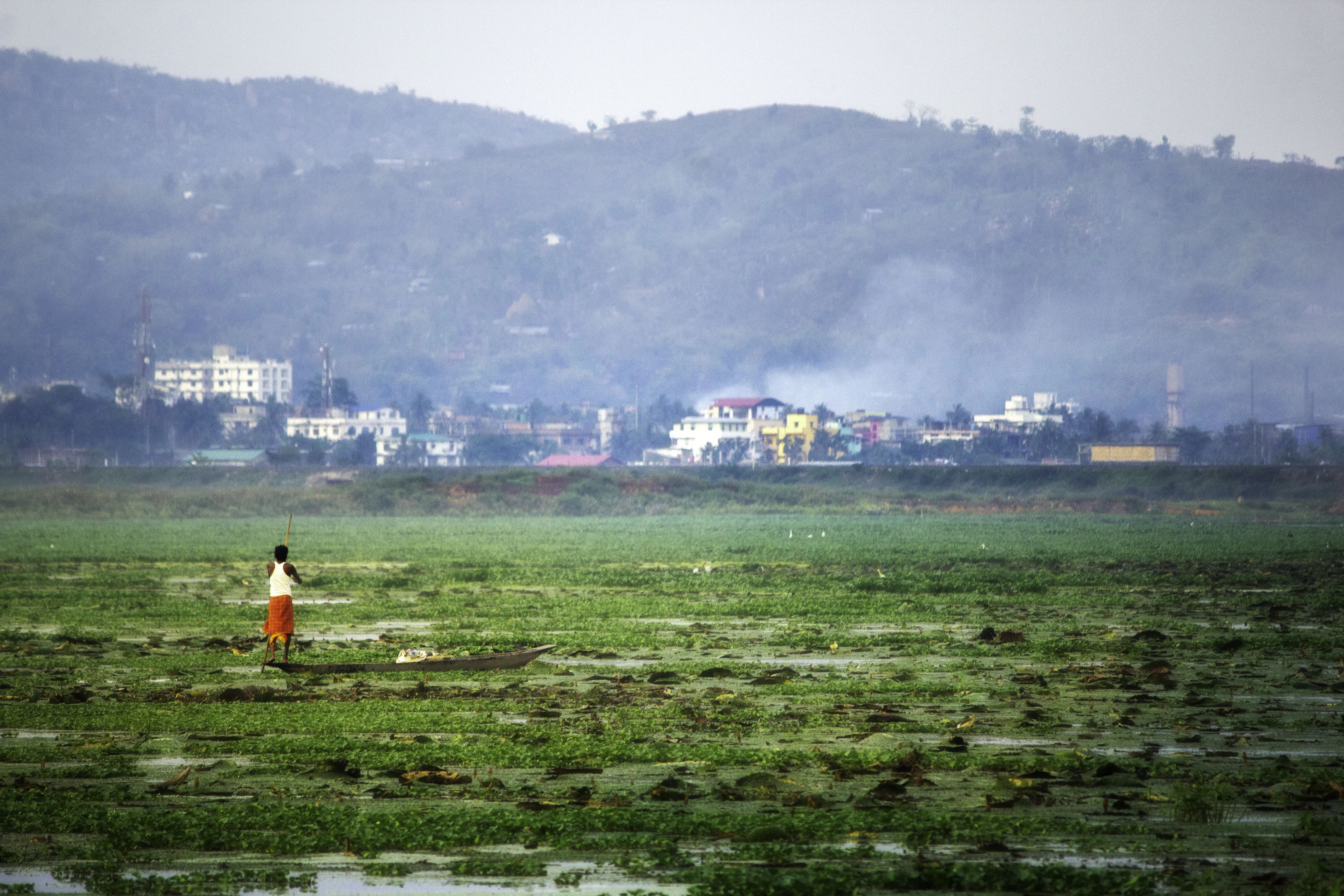 Lone figure walks across a vast green field towards buildings.